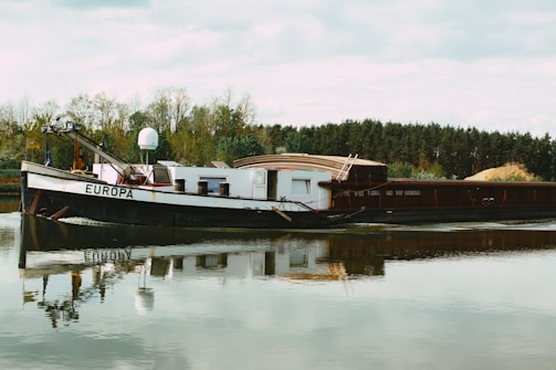 A large barge named 'Europa' is docked on a calm body of water, with its reflection visible in the water. Surrounding the barge are trees with dense foliage, suggesting a natural or rural setting. The sky is overcast, contributing to the tranquil mood.