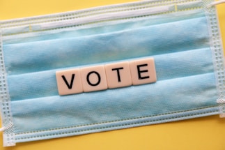 A blue surgical mask serves as the background for block letters arranged to spell 'VOTE'. The mask is placed on a yellow surface, creating a contrast with the white letter blocks.