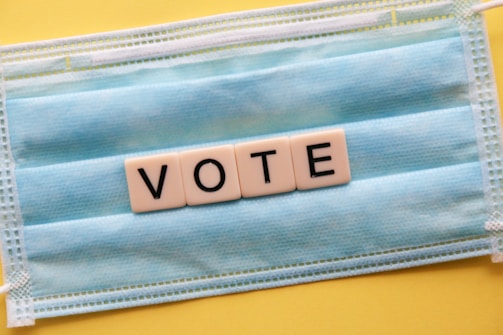 A blue surgical mask serves as the background for block letters arranged to spell 'VOTE'. The mask is placed on a yellow surface, creating a contrast with the white letter blocks.