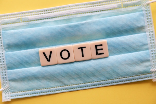 A blue surgical mask serves as the background for block letters arranged to spell 'VOTE'. The mask is placed on a yellow surface, creating a contrast with the white letter blocks.