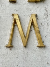 A golden letter 'M' engraved on a textured white surface. The letter appears to have some wear and dirt accumulation around its edges.