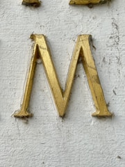 A golden letter 'M' engraved on a textured white surface. The letter appears to have some wear and dirt accumulation around its edges.