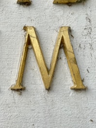 A golden letter 'M' engraved on a textured white surface. The letter appears to have some wear and dirt accumulation around its edges.