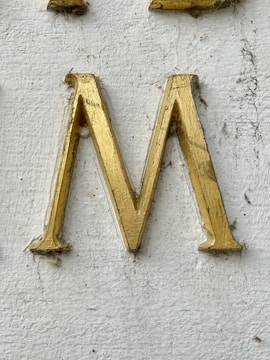 A golden letter 'M' engraved on a textured white surface. The letter appears to have some wear and dirt accumulation around its edges.