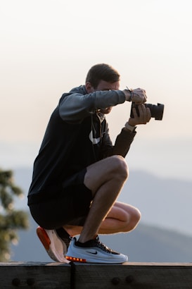 A person is crouched on a wooden platform, holding a camera up to their face, poised to take a photograph. They are wearing a dark hoodie, shorts, and white sneakers with orange highlights. The background is blurred, suggesting an outdoor setting, possibly at a vantage point with mountains visible.