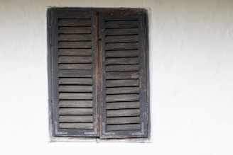 A rustic, dark wooden window with slatted shutters set against a plain off-white wall shows signs of weathering and age. The shutters are closed, and the paint is chipped, revealing the natural wood underneath.