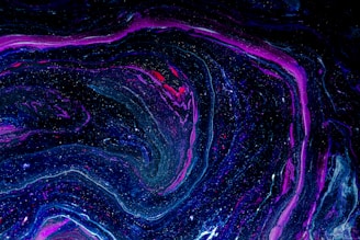 A swirling, abstract pattern featuring vivid shades of purple, blue, and black with speckles resembling stars. The flow of colors mimics a cosmic or galactic appearance, creating a sense of depth and movement.
