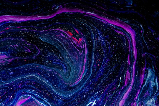 A swirling, abstract pattern featuring vivid shades of purple, blue, and black with speckles resembling stars. The flow of colors mimics a cosmic or galactic appearance, creating a sense of depth and movement.