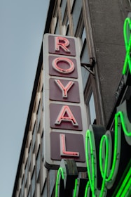 A vertical neon sign with the word 'ROYAL' in red letters displayed against a white background, attached to the facade of a building. The sign is accompanied by green neon lights underneath, and the building features multiple windows.