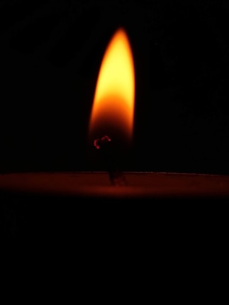 A candle flame glowing brightly against a black background. The flame displays a gradient from bright yellow and orange at the top to a darker reddish hue near the wick.