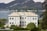 A large, elegant villa sits prominently on lush, manicured grounds with a clear view of a serene lake and towering mountains in the background. The building features classic architecture, including a symmetrical facade, tall windows, and multiple chimneys.