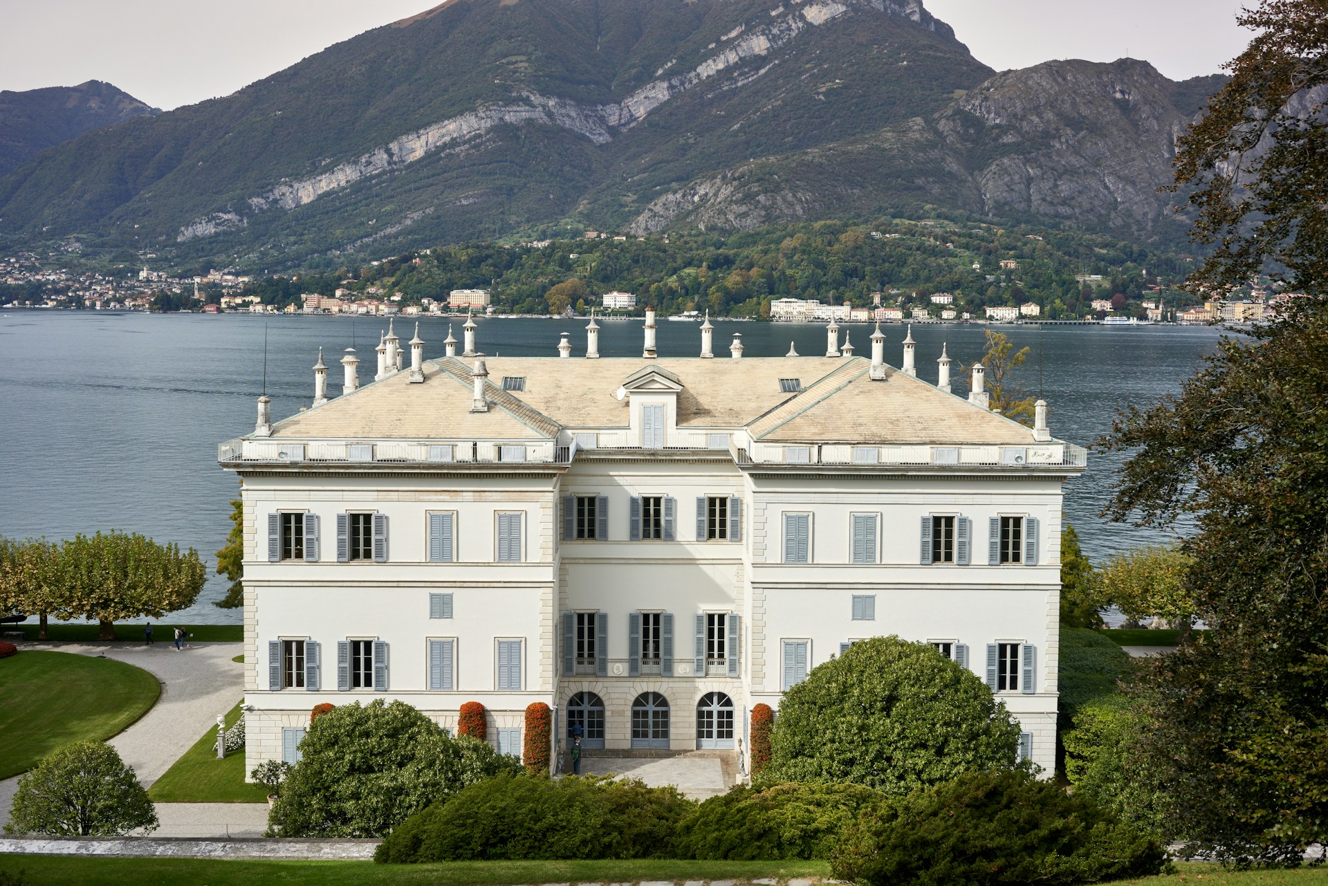 A large, elegant villa sits prominently on lush, manicured grounds with a clear view of a serene lake and towering mountains in the background. The building features classic architecture, including a symmetrical facade, tall windows, and multiple chimneys.