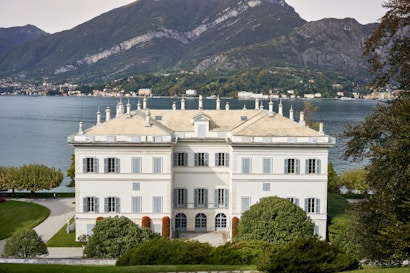 A large, elegant villa sits prominently on lush, manicured grounds with a clear view of a serene lake and towering mountains in the background. The building features classic architecture, including a symmetrical facade, tall windows, and multiple chimneys.