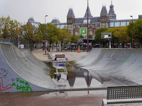 A skate park with concrete ramps partially filled with rainwater, reflecting the cloudy sky above. A shopping cart sits near the edge of a pool of water on the ramp surface. In the background, there is a grand historic building with towers, likely of European style architecture, surrounded by trees with autumn foliage. A basketball hoop stands alongside the skate park, and there is graffiti on the walls of the ramps.