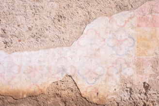 A weathered, textured wall featuring faded geometric and floral patterns in soft pastel hues. The design seems partially eroded, revealing a rough tan base underneath.