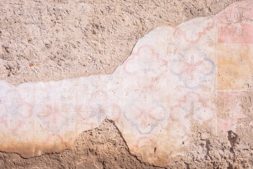 A weathered, textured wall featuring faded geometric and floral patterns in soft pastel hues. The design seems partially eroded, revealing a rough tan base underneath.