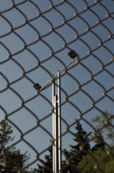 A chain-link fence dominates the foreground, while behind it, a tall metal pole with two spotlights is visible against a clear blue sky. Trees with dense foliage can be seen at the bottom of the frame, partially obscured by the fence.