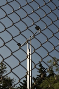 A chain-link fence dominates the foreground, while behind it, a tall metal pole with two spotlights is visible against a clear blue sky. Trees with dense foliage can be seen at the bottom of the frame, partially obscured by the fence.