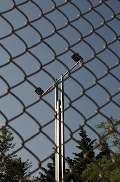 A chain-link fence dominates the foreground, while behind it, a tall metal pole with two spotlights is visible against a clear blue sky. Trees with dense foliage can be seen at the bottom of the frame, partially obscured by the fence.