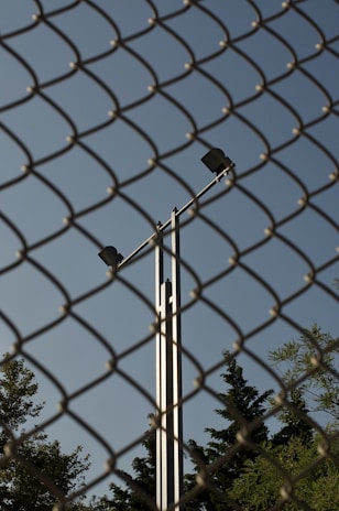 A chain-link fence dominates the foreground, while behind it, a tall metal pole with two spotlights is visible against a clear blue sky. Trees with dense foliage can be seen at the bottom of the frame, partially obscured by the fence.