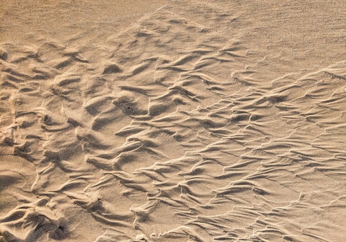 Fine sand displays intricate patterns shaped by the wind, with smooth ripples and lines creating a natural texture. The surface has varying shades from light to slightly darker sand tones.