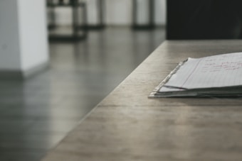 A stack of lined paper with handwritten text rests on a wooden surface. The background is out of focus, featuring a minimalist interior with grey flooring and white walls.