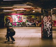 A skate park featuring colorful graffiti covering the walls and columns. There is a skateboarder in motion in the foreground, creating a sense of movement. A person stands in the background holding a skateboard under the vibrant graffiti artwork.