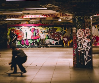 A skate park featuring colorful graffiti covering the walls and columns. There is a skateboarder in motion in the foreground, creating a sense of movement. A person stands in the background holding a skateboard under the vibrant graffiti artwork.