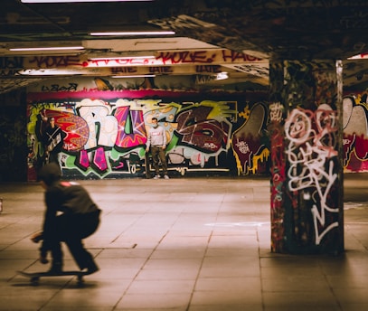 A skate park featuring colorful graffiti covering the walls and columns. There is a skateboarder in motion in the foreground, creating a sense of movement. A person stands in the background holding a skateboard under the vibrant graffiti artwork.