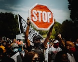 A crowd of protesters is gathered, holding various signs. One prominent sign is a modified stop sign with the words 'Stop Trivializing Our People', surrounded by names of victims. The atmosphere is intense, with people wearing masks and raising their fists or phones.