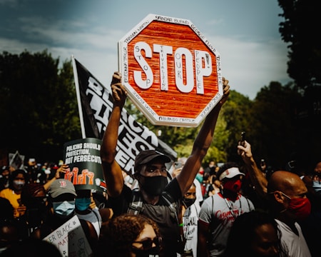 A crowd of protesters is gathered, holding various signs. One prominent sign is a modified stop sign with the words 'Stop Trivializing Our People', surrounded by names of victims. The atmosphere is intense, with people wearing masks and raising their fists or phones.