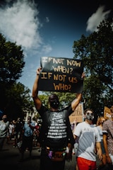 A person holds up a protest sign reading 'IF NOT NOW WHEN? IF NOT US WHO?' amidst a group of people. The scene takes place outdoors with trees in the background and a cloudy sky overhead. The crowd appears diverse, with some participants wearing masks and carrying signs expressing various messages.