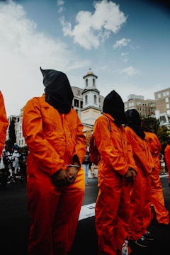Several individuals dressed in orange jumpsuits with black hoods over their heads stand in an outdoor setting, likely during a protest or demonstration. A church-like building is visible in the background, along with partially obscured city buildings and a clear sky.