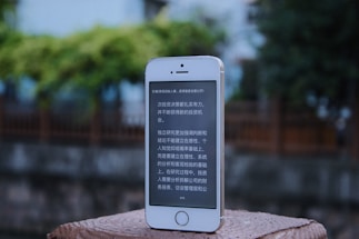A smartphone with a white and silver frame displays text in a language that resembles Chinese characters on its screen. The phone is placed on a textured, brick-like surface. In the background, there is an out-of-focus view of a wooden fence and lush greenery.