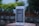 A smartphone with a white and silver frame displays text in a language that resembles Chinese characters on its screen. The phone is placed on a textured, brick-like surface. In the background, there is an out-of-focus view of a wooden fence and lush greenery.