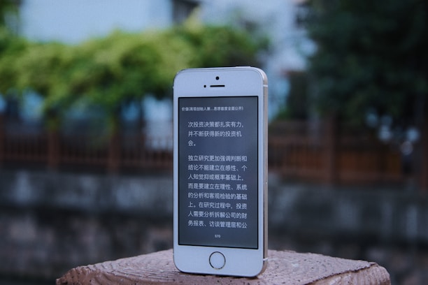 A smartphone with a white and silver frame displays text in a language that resembles Chinese characters on its screen. The phone is placed on a textured, brick-like surface. In the background, there is an out-of-focus view of a wooden fence and lush greenery.