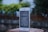 A smartphone with a white and silver frame displays text in a language that resembles Chinese characters on its screen. The phone is placed on a textured, brick-like surface. In the background, there is an out-of-focus view of a wooden fence and lush greenery.