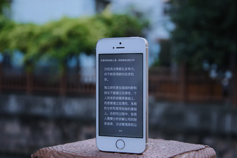 A smartphone with a white and silver frame displays text in a language that resembles Chinese characters on its screen. The phone is placed on a textured, brick-like surface. In the background, there is an out-of-focus view of a wooden fence and lush greenery.