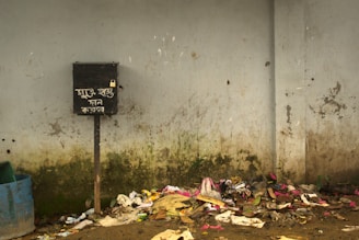 A grimy wall with visible dirt and stains, scattered litter including papers and plastic, a neglected waste area featuring a black box on a stand with foreign writing and a small padlock. An empty barrel partially enters from the left side of the image.