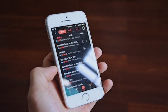 A hand is holding a white smartphone with a music playlist displayed on the screen, featuring songs by the band Pink Floyd. The background is a blurred wooden surface.