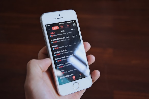 A hand is holding a white smartphone with a music playlist displayed on the screen, featuring songs by the band Pink Floyd. The background is a blurred wooden surface.