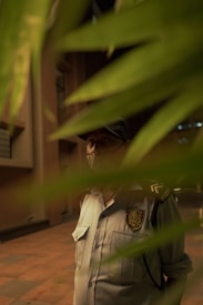 A man wearing a security guard uniform and cap stands partially hidden behind green foliage, with a visible security badge on his shirt. The background features a building with linear architectural details.