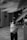 A black and white image featuring a security guard standing on a paved area of a modern building. The guard is wearing a uniform, including a shirt with shoulder insignia, a badge, a cap, and a mask. Architectural elements of the building are visible in the background, with tall geometric walls and an open passageway.