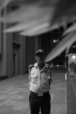 A black and white image featuring a security guard standing on a paved area of a modern building. The guard is wearing a uniform, including a shirt with shoulder insignia, a badge, a cap, and a mask. Architectural elements of the building are visible in the background, with tall geometric walls and an open passageway.