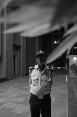 A black and white image featuring a security guard standing on a paved area of a modern building. The guard is wearing a uniform, including a shirt with shoulder insignia, a badge, a cap, and a mask. Architectural elements of the building are visible in the background, with tall geometric walls and an open passageway.