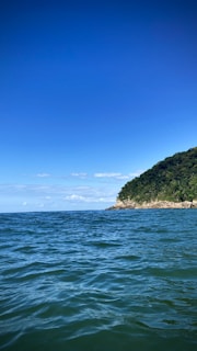A serene ocean scene with blue skies overhead. In the distance, a lush green hillside meets the water, creating a picturesque coastal view. Gentle waves ripple across the surface of the water.