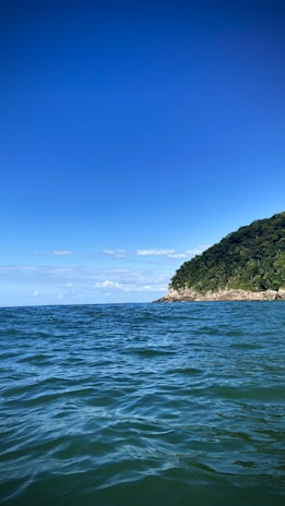 A serene ocean scene with blue skies overhead. In the distance, a lush green hillside meets the water, creating a picturesque coastal view. Gentle waves ripple across the surface of the water.