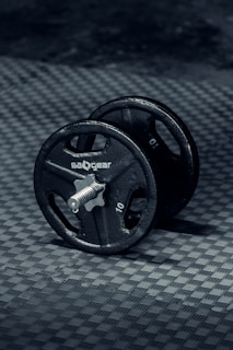A single black dumbbell is placed on a textured, dark grey surface. The dumbbell has a metallic rod with circular weights on either side, each marked with the number 10 and the brand name visible. The lighting creates subtle reflections, highlighting the metal texture and the surface beneath.