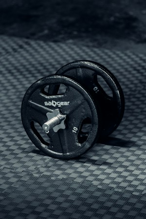 A single black dumbbell is placed on a textured, dark grey surface. The dumbbell has a metallic rod with circular weights on either side, each marked with the number 10 and the brand name visible. The lighting creates subtle reflections, highlighting the metal texture and the surface beneath.