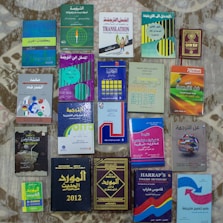 A collection of various books related to translation and dictionaries, with covers in a mix of English and Arabic languages. The books have diverse cover designs featuring text, globes, and other graphical elements. They're arranged in neat rows on a patterned surface.
